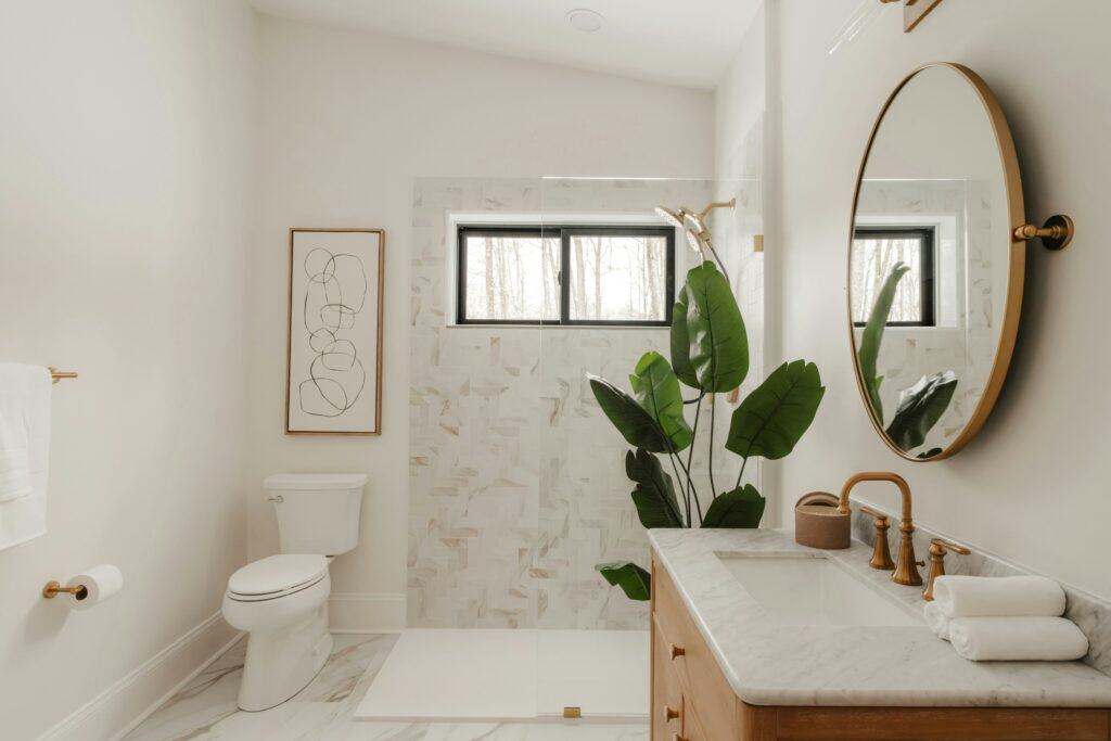 White bathroom with a round gold-framed mirror, marble-tiled shower and a large leafy plant
